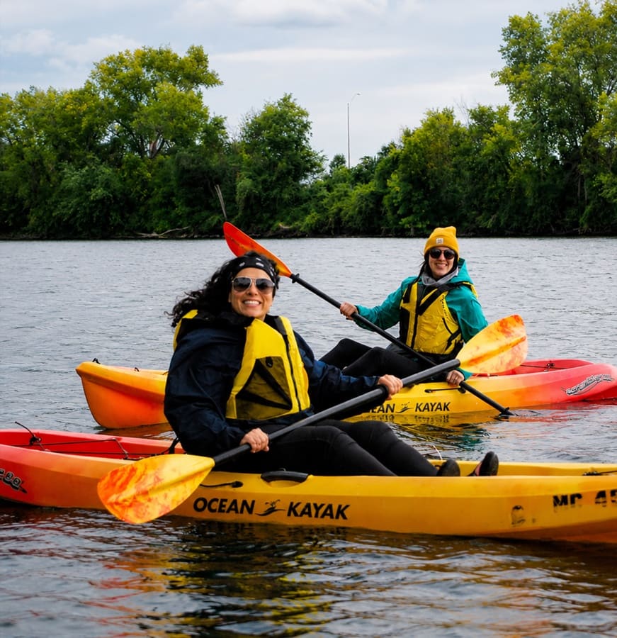 Two women smiling as they paddle bright kayaks on calm coastal water