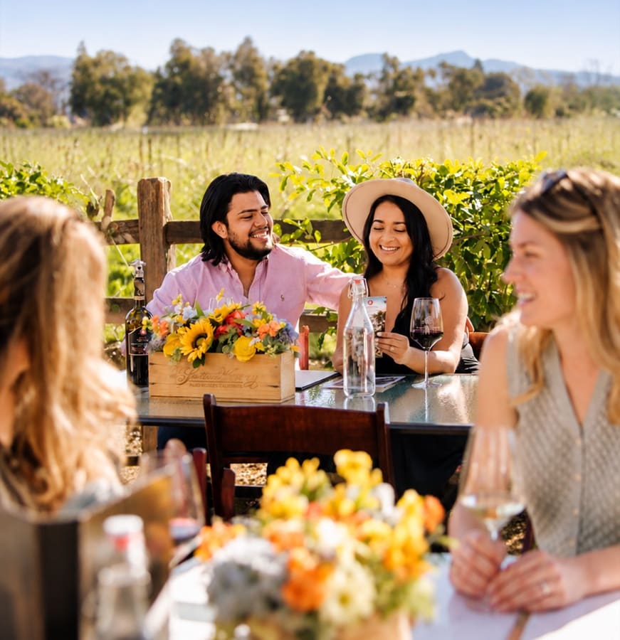 Friends laughing over glasses of wine at an outdoor table in a vineyard